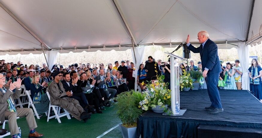 Joe Erwin, chairman and owner of Greenville Triumph SC and Greenville Liberty SC, addressed an enthusiastic crowd of the groundbreaking in Mauldin. (Photo/Sarah Marko)|Ronaldinho took the lead on shoveling the ceremonial ground at the future site of Triumph Stadium. (Photo/Sarah Marko)|Fans of Ronaldinho took turns posing for photos in the seat he occupied during the ceremony. (Photo/Ross Norton)|Triumph Stadium will be constructed in a now vacant lot next to BridgeWay Station's shops and eateries. (Photo/Ross Norton)