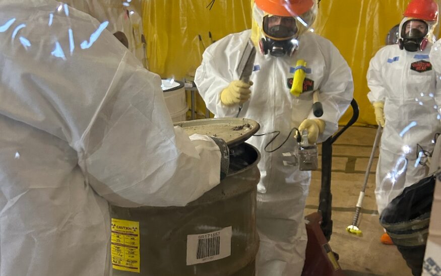 Operators at the Solid Waste Management Facility at the Savannah River Site are removing waste drums from overpacks to separate drums ready to be shipped to the Waste Isolation Pilot Plant from those that are not yet ready for shipment. The overpacks are standard waste boxes containing a group of drums. They perform removal activities in a plastic hut, using full personal protective equipment. (Photo/Savannah River Nuclear Site)